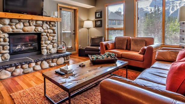 A cozy living room with a stone fireplace, leather sofa, and large windows. - Spring Creek Vacations 2 BR Premier Condo 115R (Canmore)