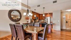 A spacious dining area with a wooden table and leather chairs, a kitchen with modern appliances, and a large mirror on the wall. - Spring Creek Vacations 2 BR Premier Condo 115R (Canmore)