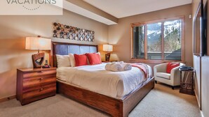 A hotel room with a large bed, bedside lamps, a wooden nightstand, a chair, and a window with blinds. - Spring Creek Vacations 2 BR Premier Condo 115R (Canmore)