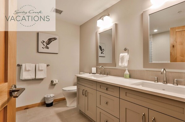 A modern bathroom with a large mirror, two sinks, and a toilet. There are towels hanging on the wall and a trash can near the toilet.