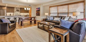 A spacious living area with a sofa, coffee table, and a dining table. There is a kitchen in the background with modern appliances and wooden cabinets. - Spring Creek Vacations 3 BR Premier Condo 103W (Canmore)
