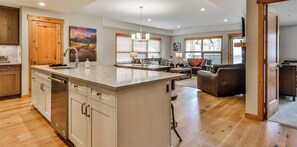 A spacious kitchen with a large island, wooden cabinets, and a dishwasher. There is a dining area with a table and chairs, and a living room with a sofa and a TV. - Spring Creek Vacations 3 BR Premier Condo 103W (Canmore)