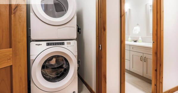 Zona de lavandería con dos lavadoras Whirlpool apiladas, puertas de madera y un vistazo a un baño con espejo y artículos de aseo.