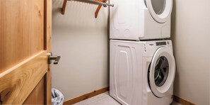 A laundry area with a stacked washer and dryer, a wooden drying rack, and a door to a room.