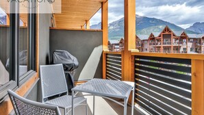 A balcony with metal chairs and a barbecue grill, overlooking a row of wooden cabins with a mountainous backdrop.