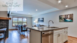 A spacious kitchen with a central island, a dining area with chairs, a sofa, and a fireplace. There is a large window providing natural light and a painting on the wall.