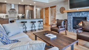 A modern living room with a fireplace, a TV mounted above it, and a kitchen area with wooden cabinets and a stainless steel refrigerator.
