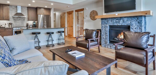 A modern living room with a fireplace, a TV mounted above it, and a kitchen area with wooden cabinets and a stainless steel refrigerator.