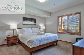 A bedroom with a large bed, two bedside tables with lamps, a painting on the wall, and a window with blinds.