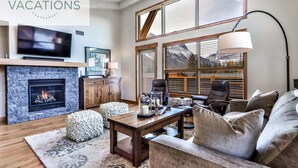 A modern living room with a fireplace, comfortable seating, a television, and large windows offering a view of the outdoors. - Spring Creek Vacations 3 BR Premier Condo 309W (Canmore)