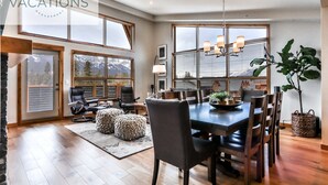A spacious dining area with a large wooden table, multiple chairs, and a chandelier. There are large windows providing views of the outdoors. - Spring Creek Vacations 3 BR Premier Condo 309W (Canmore)