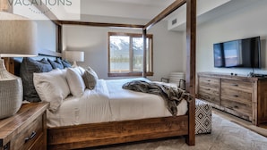 A spacious bedroom with a large bed, wooden headboard, bedside tables, a TV, and a window with blinds. - Spring Creek Vacations 3 BR Premier Condo 309W (Canmore)