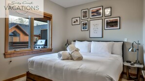 Een net opgemaakt bed met witte lakens en handdoeken, een bedlamp en ingelijste foto's aan de muur.