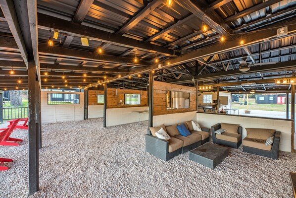 A spacious indoor area with a high ceiling, wooden beams, and string lights. There are seating areas with cushions and a gravel floor.
