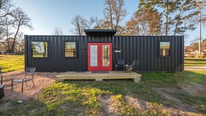 A modern, compact cabin with a red door, two windows, and a small porch with chairs. - Hawg Boss: 2 mins to Lake Conroe! Heated Pool, Fire Pit, Stocked Ponds (Willis)
