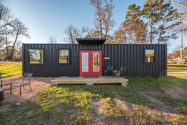 Une cabane moderne et compacte, dotée d’une porte rouge, de deux fenêtres et d’un petit porche avec des chaises.
