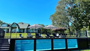 A swimming pool area with lounge chairs and umbrellas, surrounded by a fence and houses in the background. - Charcoal King: 2 mins to Lake Conroe! Heated Pool, Fire Pit, Stocked Ponds (Willis)
