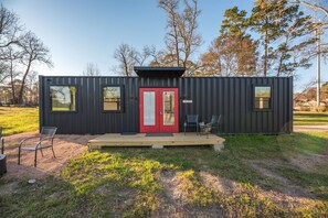 Une cabane moderne et compacte, dotée d’une porte rouge, de deux fenêtres et d’un petit porche avec des chaises.
