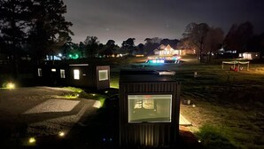 En rad små stugor med stora fönster, en väl upplyst gångväg och ett hus i fjärran med upplysta fönster.