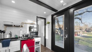 Una cocina moderna con armarios blancos, un refrigerador rojo y un microondas. Hay una ventana con persianas y vistas a un patio trasero con una hamaca y un árbol.
