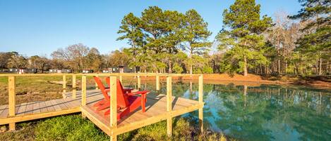 Un muelle de madera con sillas rojas que se extiende sobre un lago tranquilo, rodeado de árboles y un cielo despejado.