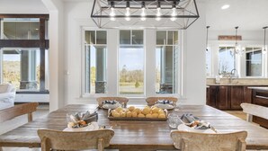 A spacious dining area with a long wooden table set for six, large windows offering a view of the outdoors, and a modern kitchen with dark wood cabinets and a stone countertop. - LUXURY home 2 mins to Lake Conroe! Heated Pool, Stocked Ponds, Cute Pygmy Goats (Willis)