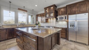 A spacious kitchen with dark wood cabinets, a large island with a granite countertop, stainless steel appliances, and a view of the outdoors through large windows. - LUXURY home 2 mins to Lake Conroe! Heated Pool, Stocked Ponds, Cute Pygmy Goats (Willis)