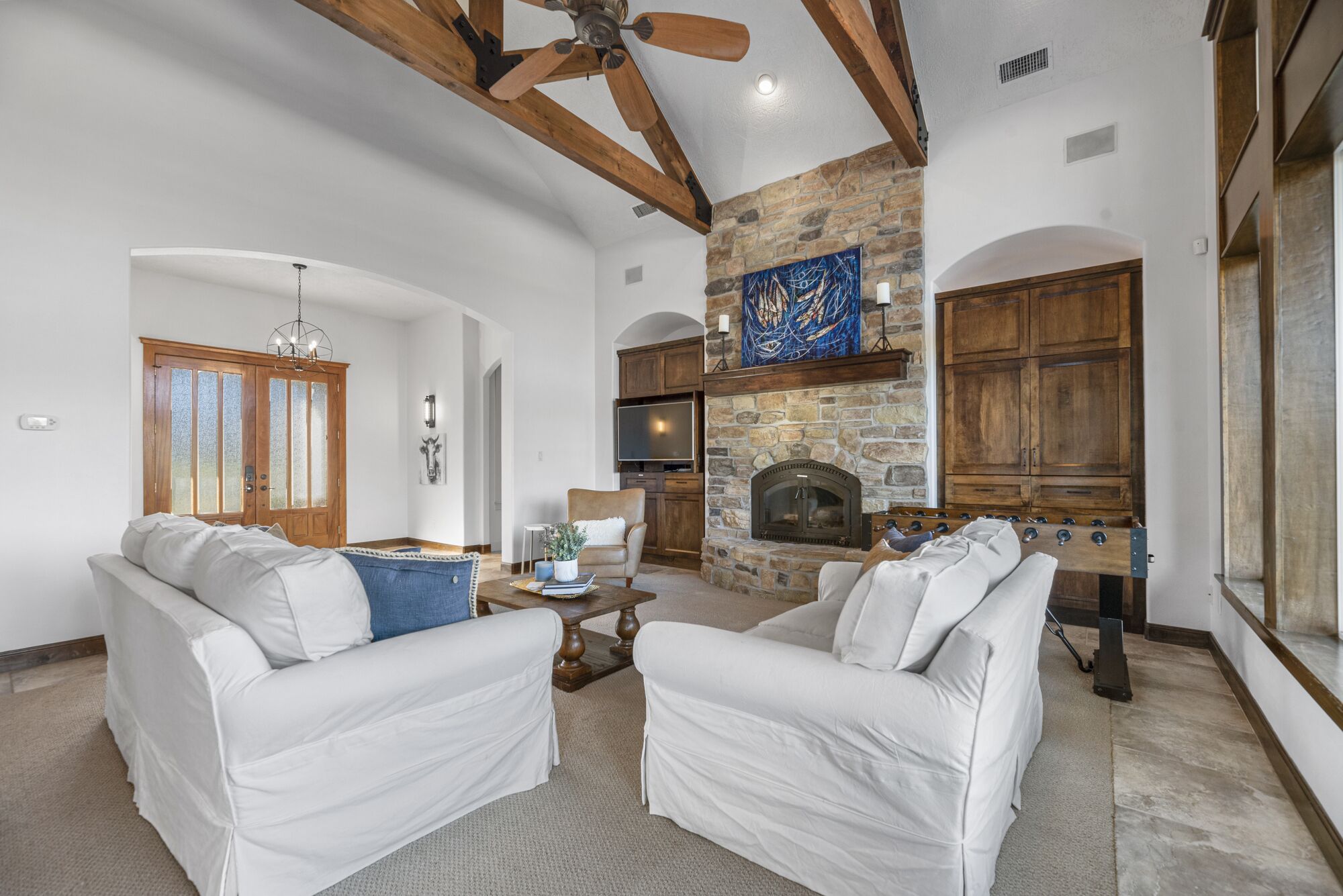 A spacious living room with white sofas, a stone fireplace, wooden beams, and a television.