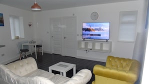 Un salon avec un canapé blanc, un fauteuil jaune, un téléviseur à écran plat, une table à manger avec des chaises et une petite table blanche.