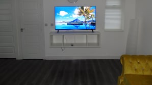 Un salon avec un téléviseur à écran plat posé sur une étagère blanche, une horloge au mur et un fauteuil jaune.