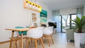 Ein modernes Wohnzimmer mit Essbereich, ausgestattet mit einem Holztisch, weißen Stühlen, einem Fernseher und einem großen Fenster mit Vorhängen.