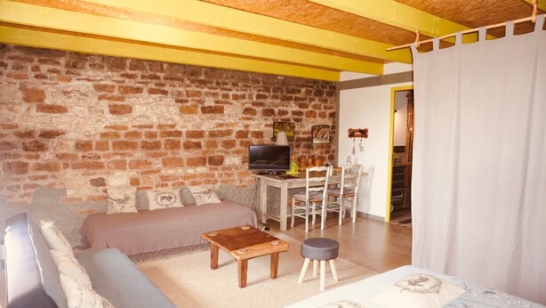 A cozy living room with a sofa, a coffee table, and a TV. There is a dining area with chairs and a table. The room has a brick wall and wooden beams on the ceiling.