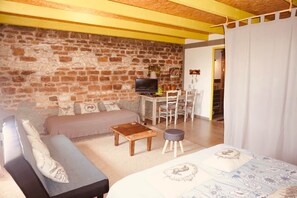 Een gezellige woonkamer met een bank, een salontafel en een televisie. Er is een eethoek met stoelen en een tafel. De kamer heeft een bakstenen muur en houten balken aan het plafond.