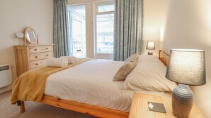 A neatly made bed with a wooden headboard, a bedside table with a lamp, a dresser with a mirror, and a window with curtains. - 48 Glan Y Mor (Fairbourne)