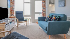 A modern living room with a blue sofa, a blue armchair, and a TV on a wooden desk. - 48 Glan Y Mor (Fairbourne)