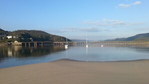 A calm waterfront with a bridge, boats, and a sandy beach. - 48 Glan Y Mor (Fairbourne)