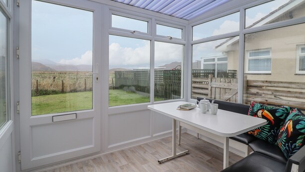 A conservatory with a view of a grassy field and mountains. It includes a table with a chair and a bench. - 48 Glan Y Mor (Fairbourne)