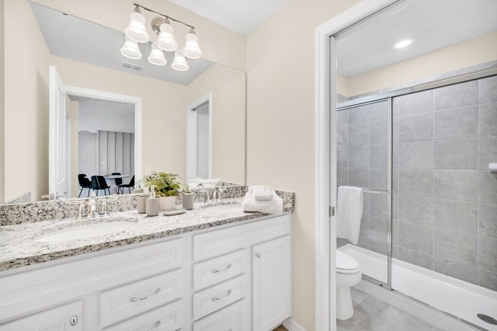 A modern bathroom with a large mirror, white cabinetry, a granite countertop, and a glass shower enclosure.