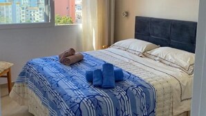 A bedroom with a bed, a window with a view of buildings, a ceiling fan, and a painting on the wall. - Beautiful apartment with pool (Torremolinos)