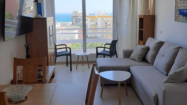 A modern living room with a sofa, a dining table, and chairs. There is a TV mounted on the wall, a painting, and a view of the city through large windows. - Beautiful apartment with pool (Torremolinos)