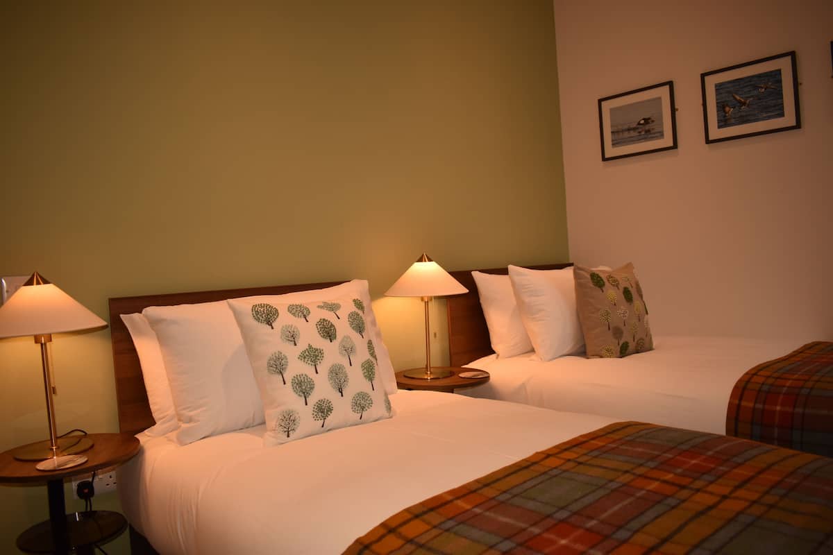 A hotel room with two beds, each with a lamp, a plaid blanket, and a wall with two framed pictures.