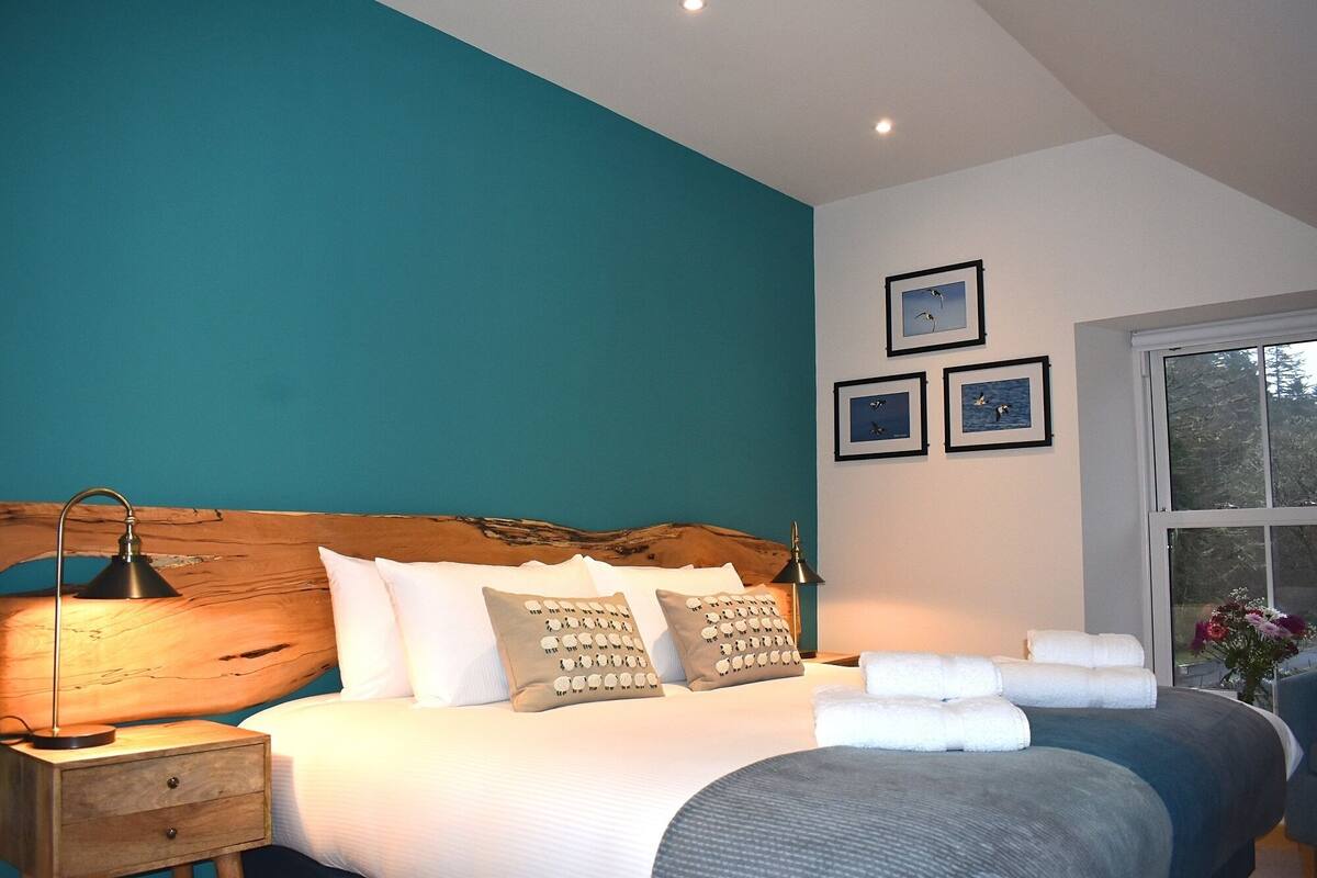 A bedroom with a wooden headboard, a large bed with white bedding and patterned pillows, a bedside table with a lamp, and a window with a view of trees.