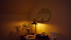 Ein schwach beleuchteter Raum mit Schreibtisch, Lampe, Spiegel, einer Vase mit Blumen, einem Buch, einer Kaffeemaschine und einer Tasse.
