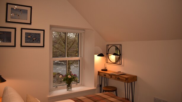 Ein Schlafzimmer mit einem Bett, einem Schreibtisch, einem Stuhl, einem Fenster mit Blick auf Bäume und gerahmten Bildern an der Wand.