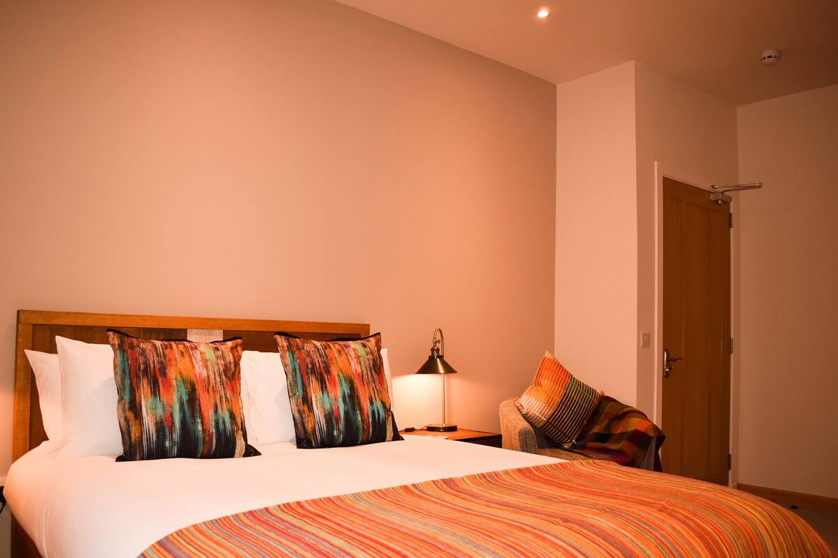 A hotel room with a wooden headboard, a bed with white linens, colorful striped bedding, a bedside lamp, and a door to the right.