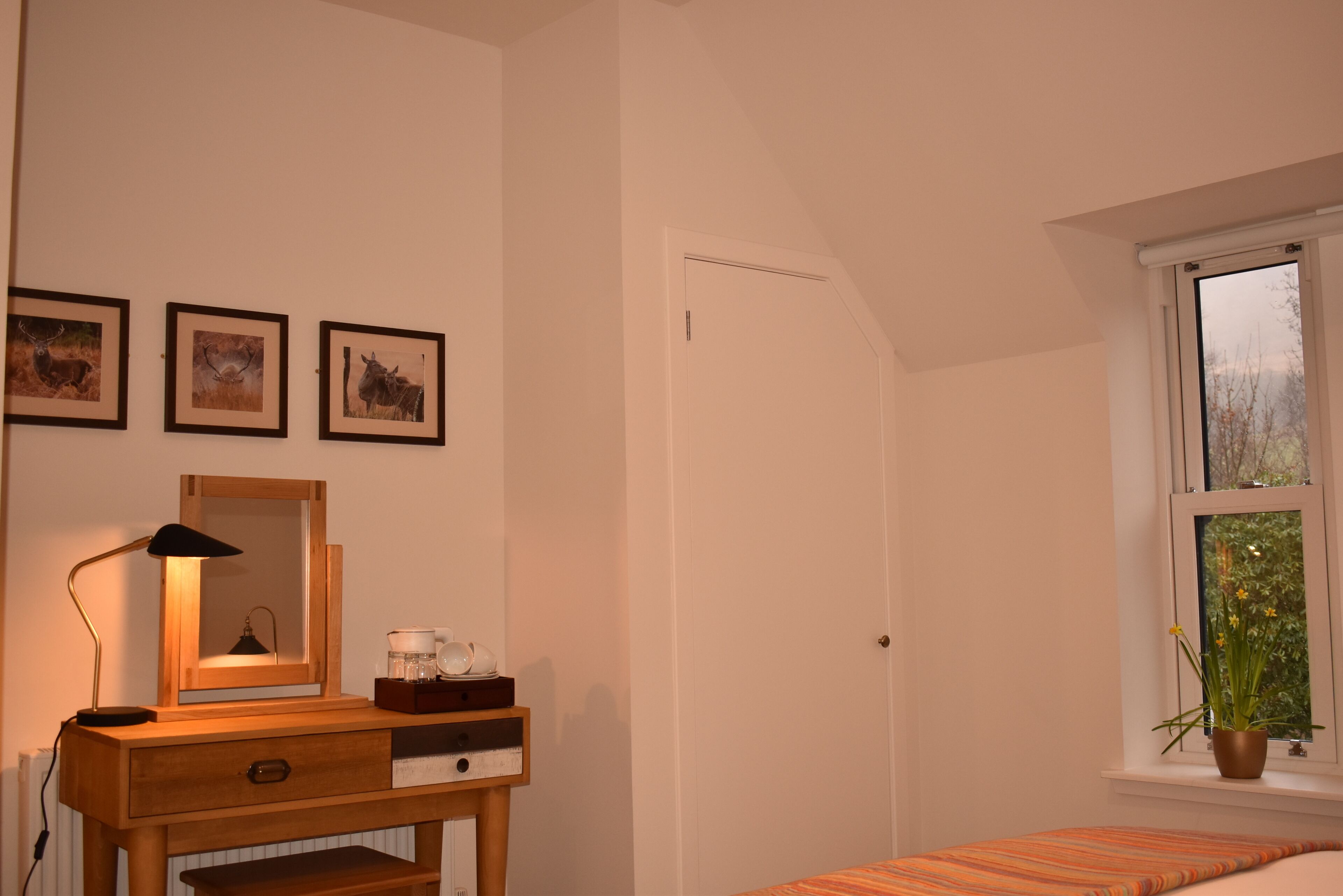 A bedroom with a wooden desk, a mirror, framed pictures on the wall, a window with a view of greenery, and a bed with a striped bedspread.