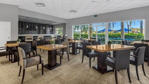 Una sala de conferencias moderna con varias mesas y sillas, ventanas amplias con vistas a una piscina y palmeras, y una zona de kitchenette.