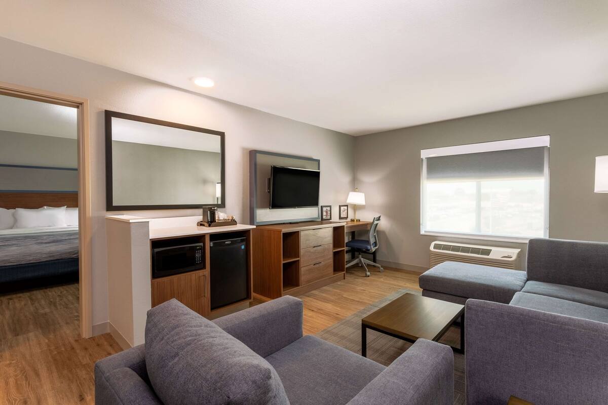 A compact hotel room with a kitchenette, a flat-screen TV, a sofa, and a large window providing natural light.