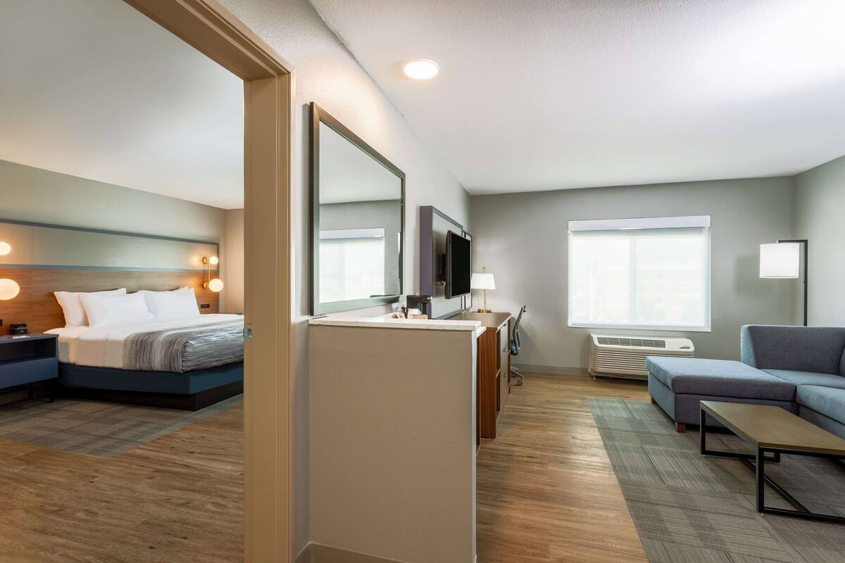 A modern hotel room with a bed, a desk, a TV, a sofa, and a large window.