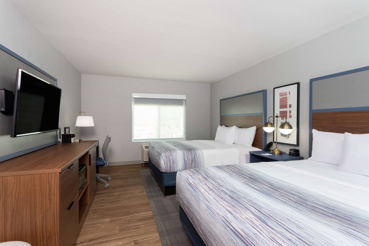 A hotel room with a flat-screen TV, two beds with striped bedding, a desk with a lamp, a window, and a headboard with a mirror and lamps.